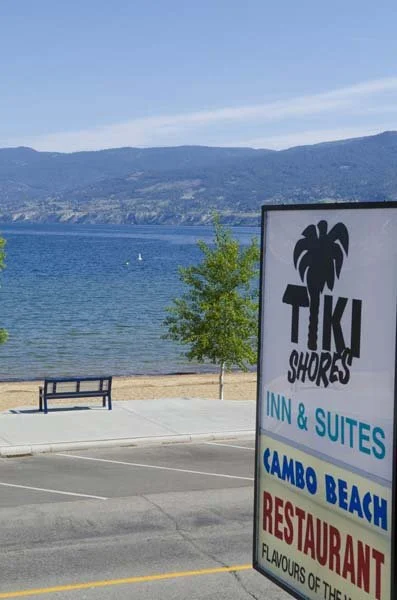 Sign for Tiki Shores Inn & Suites near a beach with water and mountains in the background.