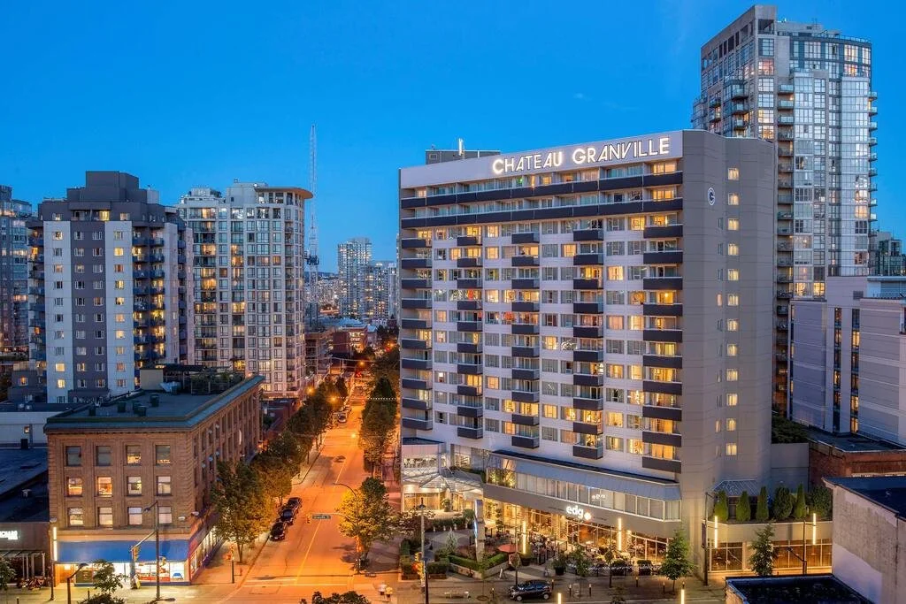 Tall modern hotel building labeled 'Chateau Granville' in a cityscape during twilight, surrounded by high-rise apartments and a tree-lined street with some cars and pedestrians.