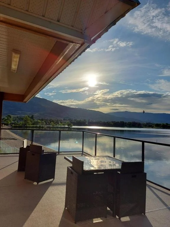 Outdoor patio with wicker furniture and a glass table overlooking a lake with mountains in the background under a partly cloudy sky with the sun shining.