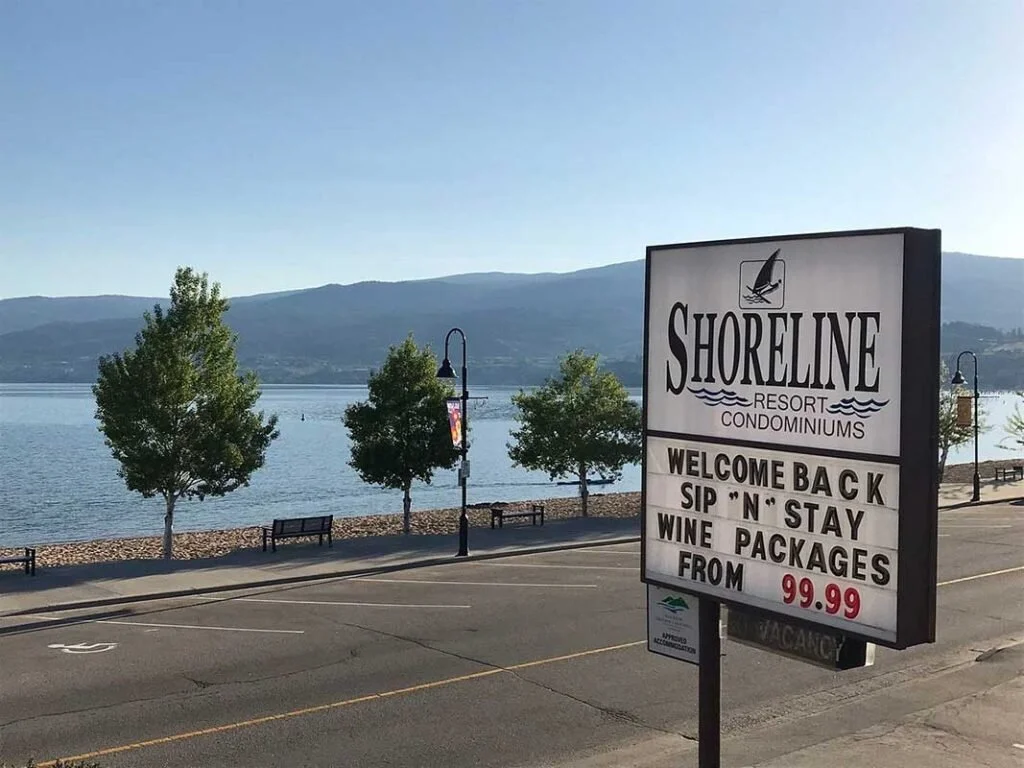 View of a lakeside promenade with trees, benches, and street lamps, and a large sign advertising Shoreline Resort Condominiums and wine packages starting at $99.99, with mountains in the background.