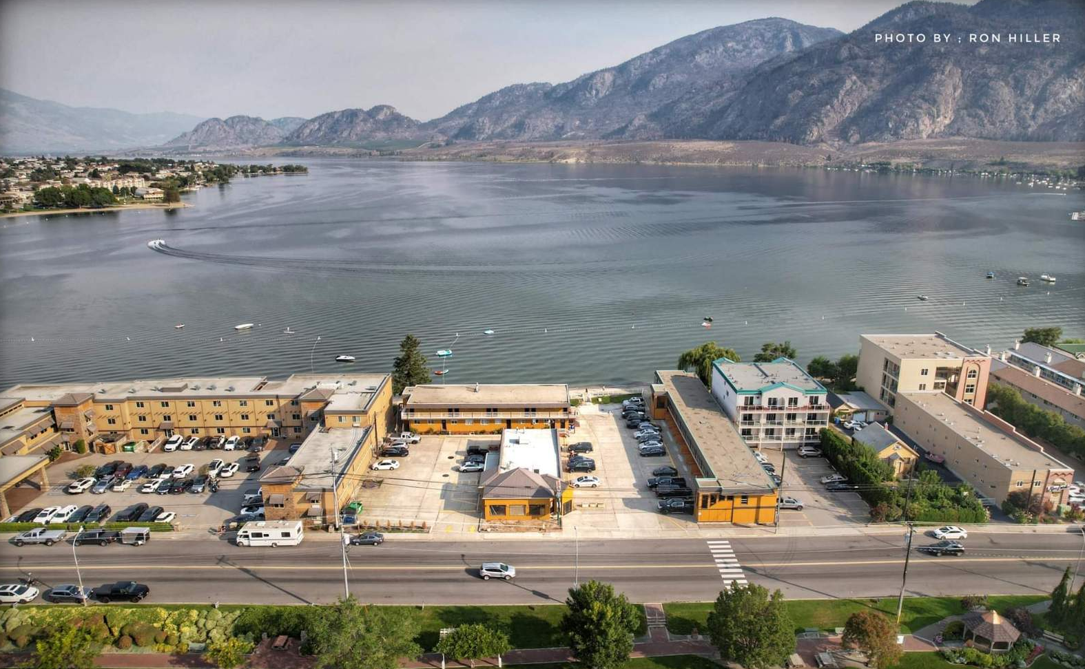Aerial view of a lakeside town with apartment buildings, parked cars, and boats on the water, mountains in the background.