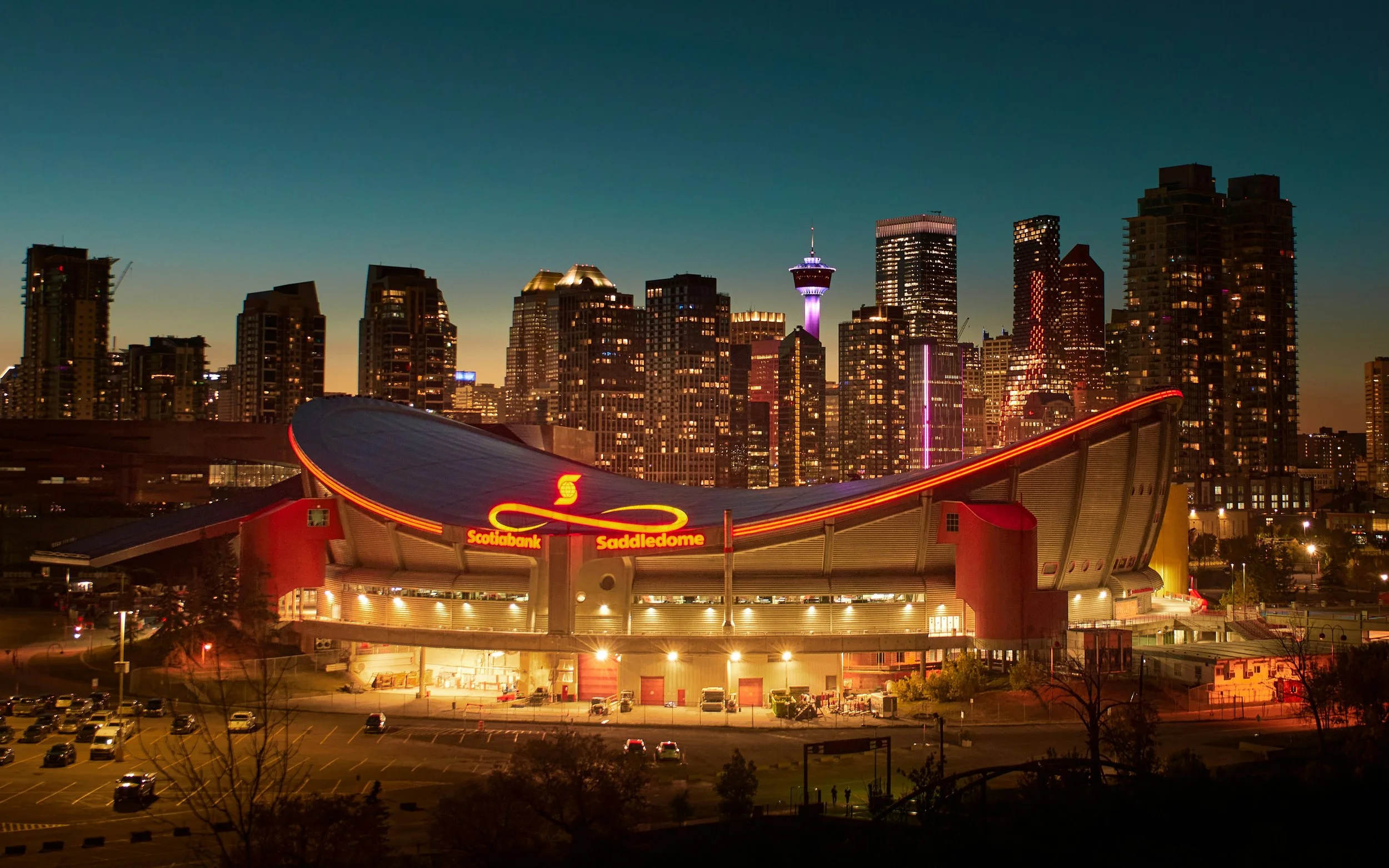 Nighttime city skyline with tall buildings, a tower lit with purple lights, and a sports stadium with a red sign reading 'Scotiabank Saddledome' in the foreground.