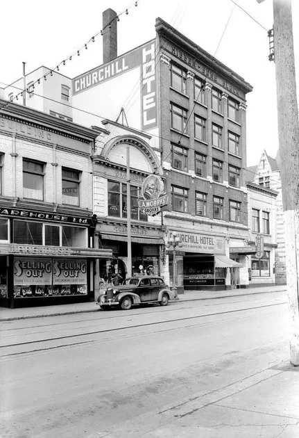 Black and white photo of a city street featuring the Churchill Hotel building with multiple signs, including a large sign for the hotel and a marquee for a theater or business, a vintage car parked on the street, and storefronts with 