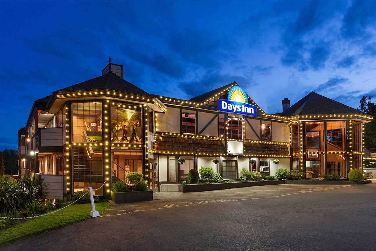 A two-story Days Inn hotel building decorated with string lights during the evening, with large windows showing the interior, and an empty parking lot in front.