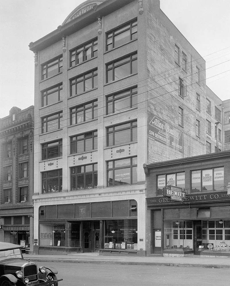 Black and white photo of a multi-story commercial building with large windows on each floor, located on a city street. The ground floor has a storefront with a sign that reads "Insurance." Adjacent storefronts display signs for products like badges, stamps, and rubber stamps. An older car is parked in front of the building.