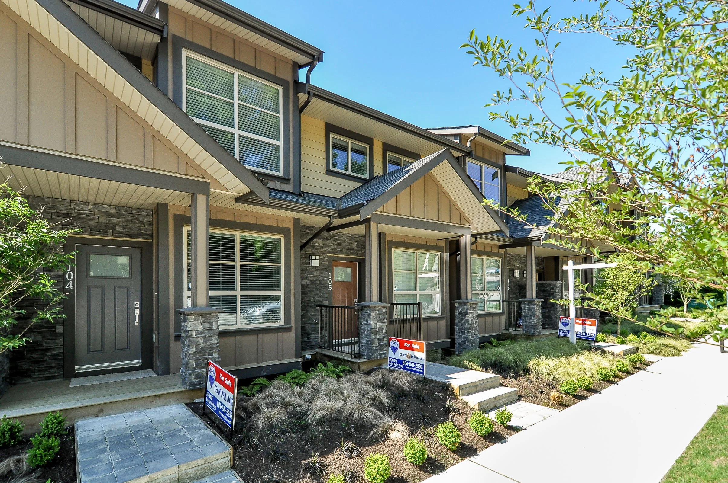 A row of modern townhouses with brick and siding exteriors, small front porches, and 'For Sale' signs in front yards, under a clear blue sky.