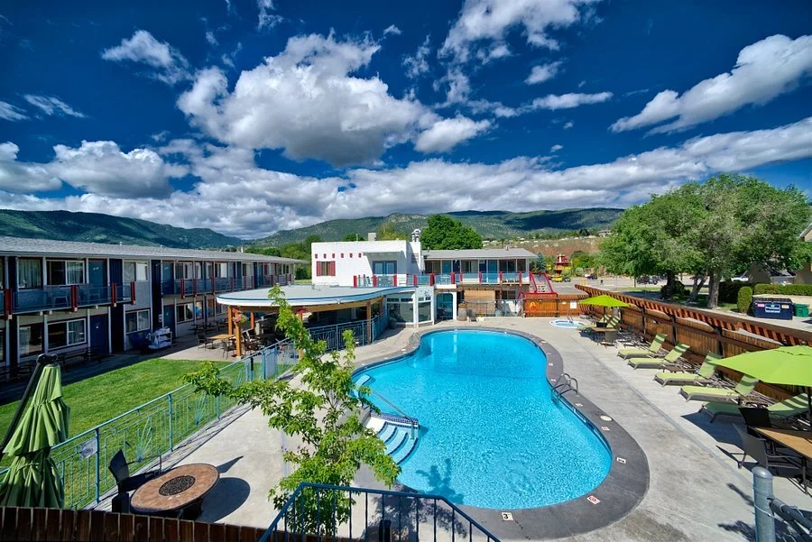 Outdoor swimming pool with lounge chairs and umbrellas at a hotel or resort, with apartment buildings and mountains in the background under a partly cloudy sky.