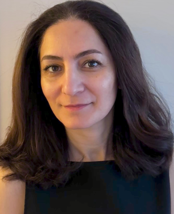 A woman with long, dark brown hair and light skin, wearing a black top, smiling slightly against a neutral background.