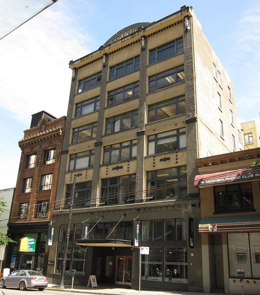 A multi-story building with large windows and a distinctive facade, located on an urban street with parked cars and nearby shops.
