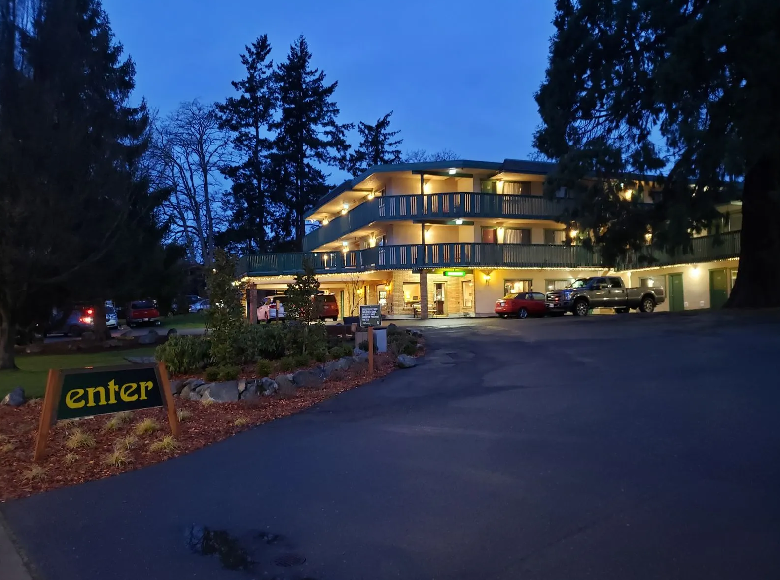 A multi-story hotel at dusk with exterior lights, surrounded by trees and parked cars, and a sign that reads 'enter' at the entrance.