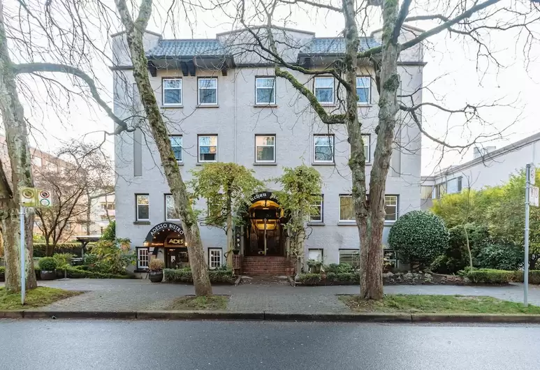 Gray multi-story heritage hotel with a restaurant entrance named 'Adesso Bistro,' surrounded by trees and greenery, located on a street with a pedestrian sidewalk.