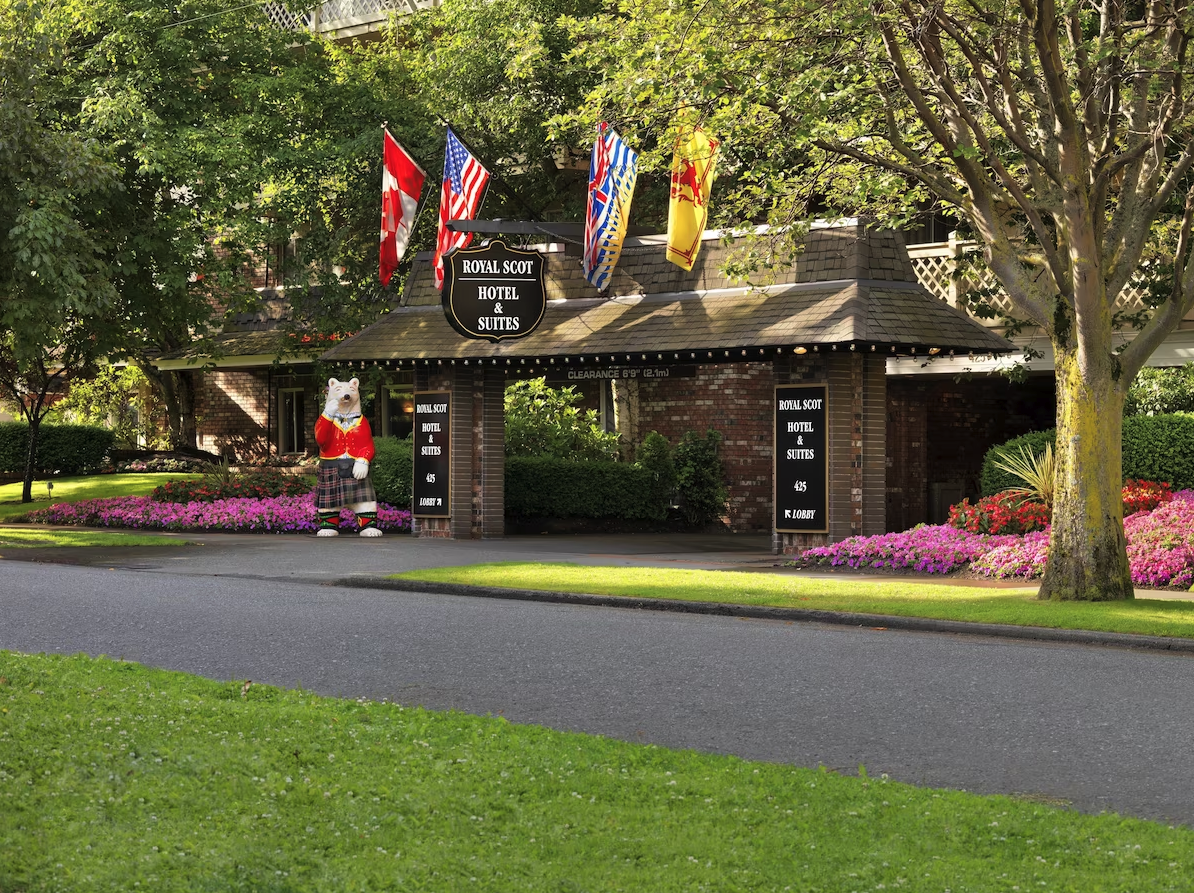 Entrance to the Royal Scot Hotel & Suites with flags, a bear statue dressed in Scottish attire, and colorful flower beds.