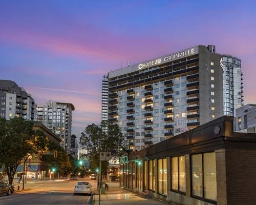 A cityscape at dusk featuring the Chateau Granville hotel, high-rise buildings, a street with cars, and a pink-purple sky.