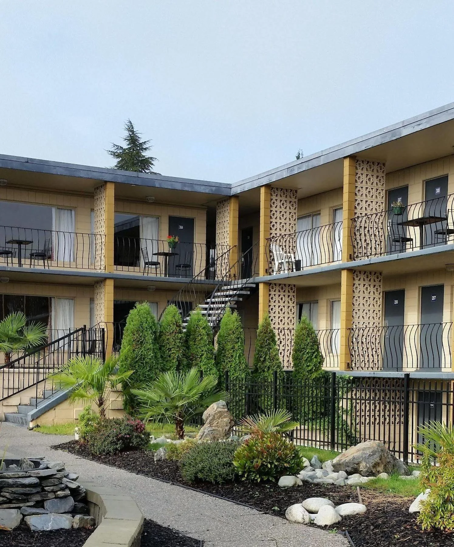 A multi-story motel with yellow walls and black railings on the balconies, surrounded by a landscaped garden with various shrubs, rocks, and palm trees.