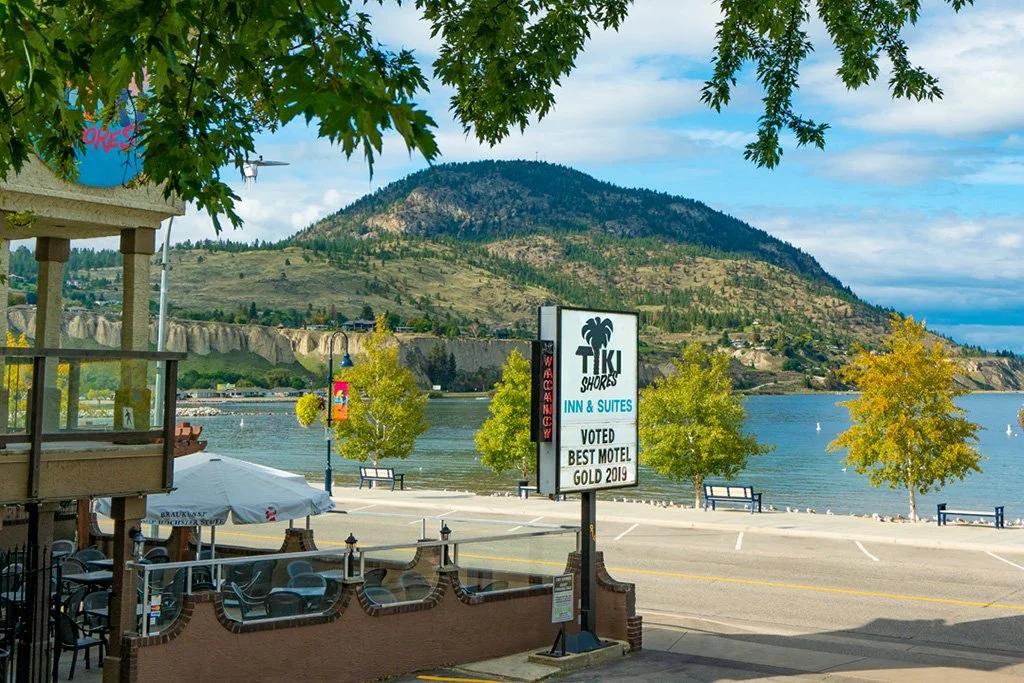 View of a small lakeside town with a mountain in the background, trees with green and yellow leaves, a sign for Tiki Shores Inn & Suites, and outdoor seating under umbrellas.