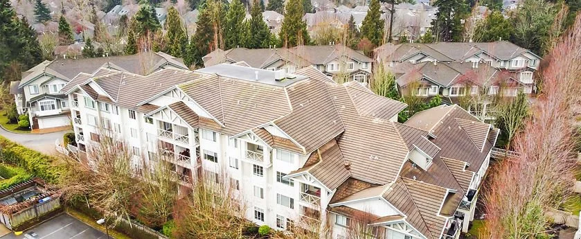 An aerial view of a residential neighborhood with multi-story apartment buildings, brown tiled roofs, and surrounded by trees and other houses.