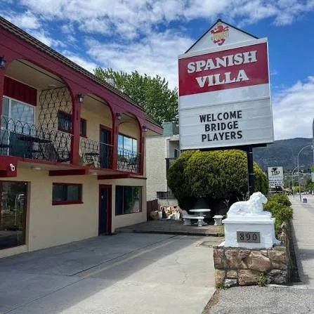 Sign for Spanish Villa restaurant welcoming bridge players, with a building featuring a red balcony and a large bush in front.