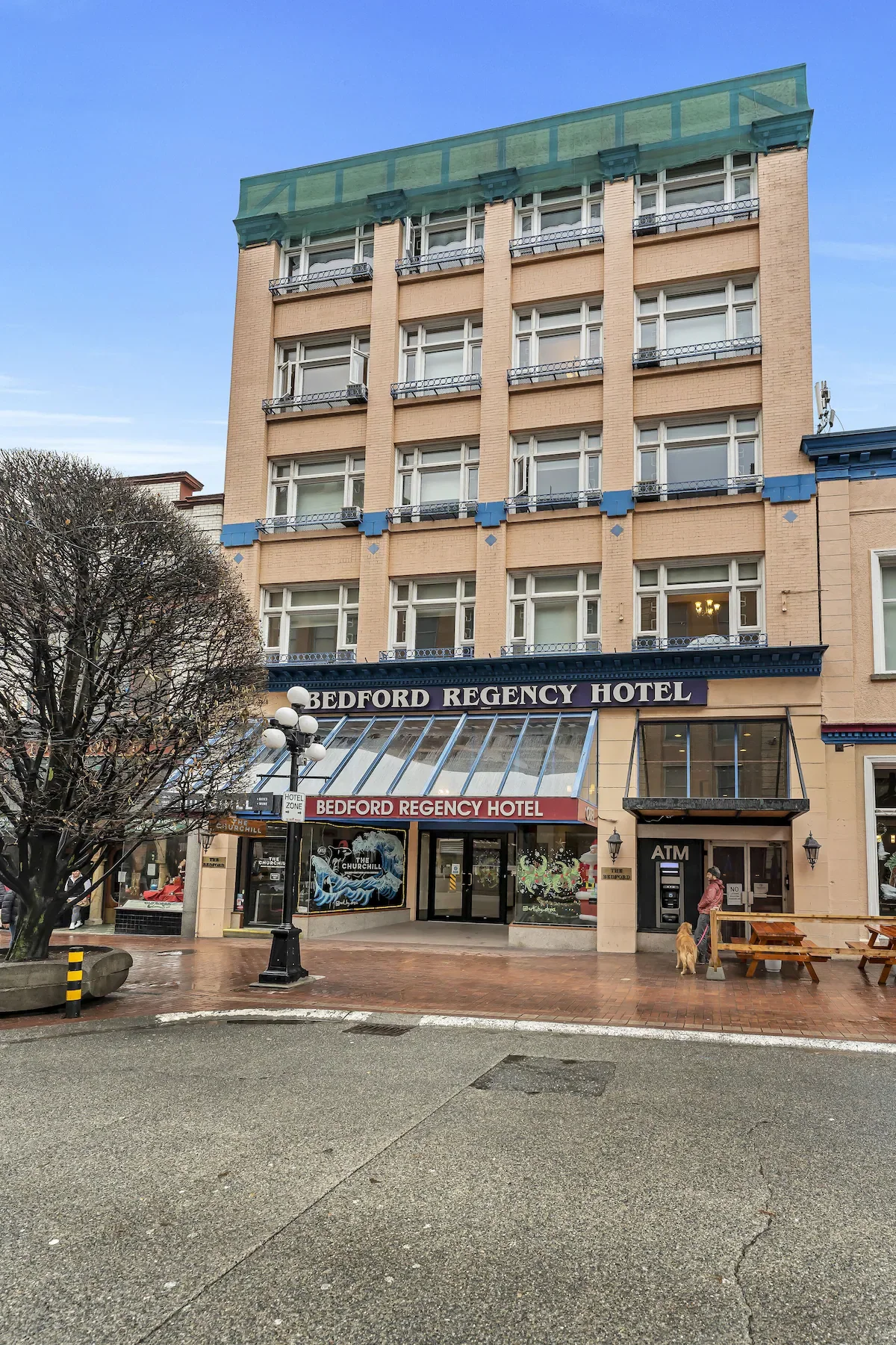 The Bedford Regency Hotel building with a glass awning and a sign. The street scene shows a person with a dog near a bench and an ATM, with a tree and lamppost in the foreground.