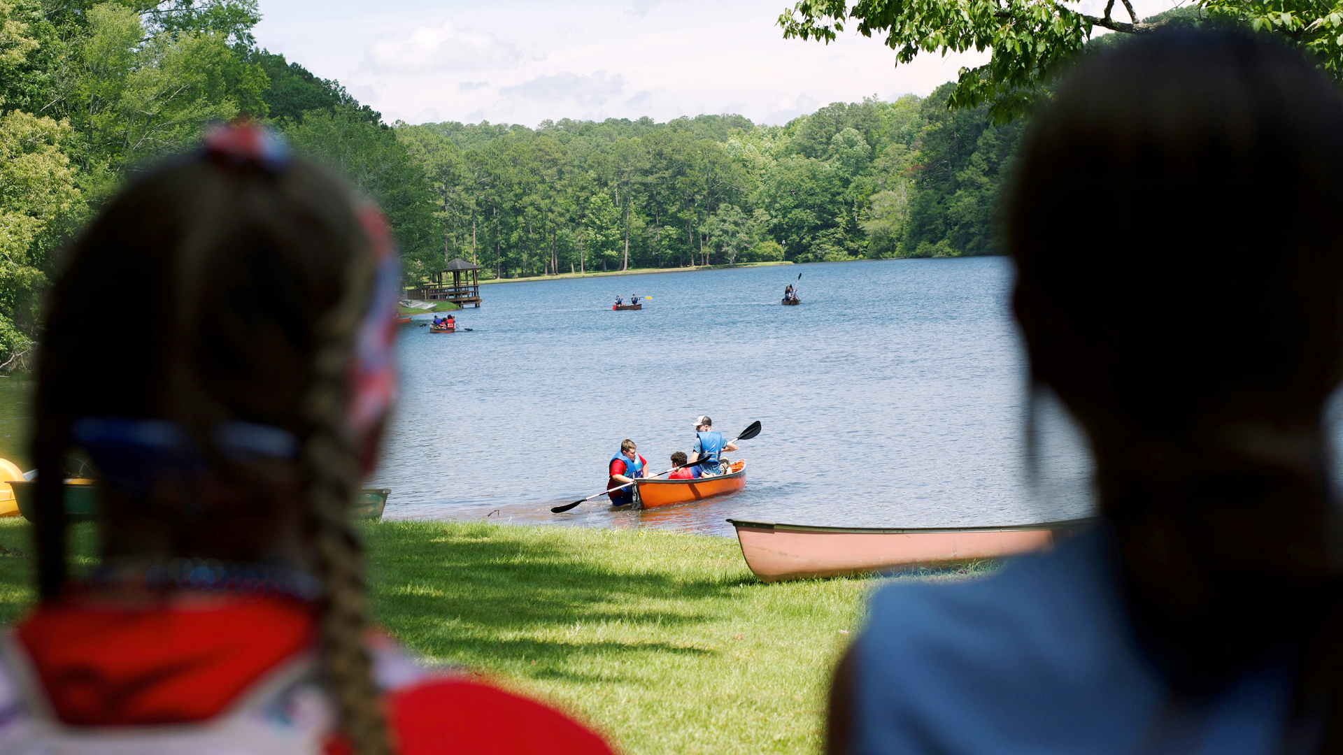 People kayaking on a lake, viewed through the silhouettes of two children in the foreground, with a lush, green forest in the background.