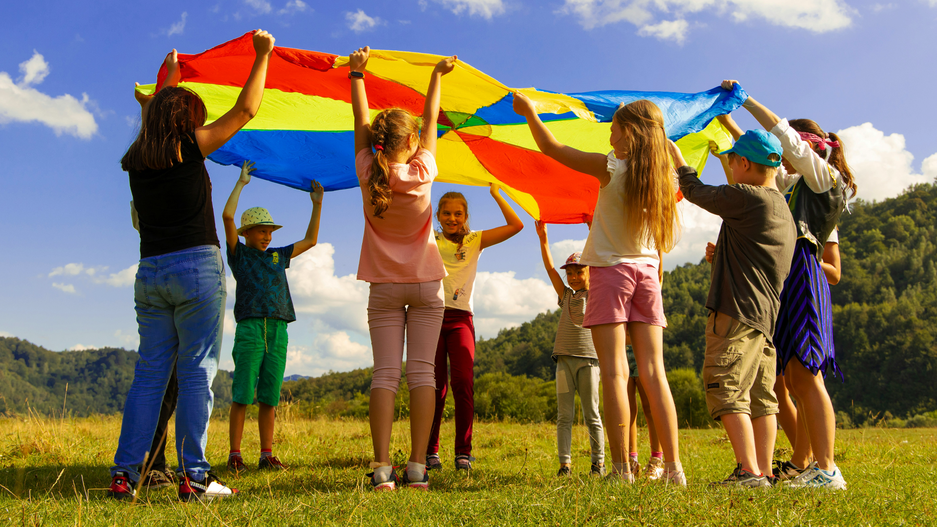 Children playing outdoors with a colorful parachute in a grassy field, with a backdrop of trees and a blue sky with clouds.