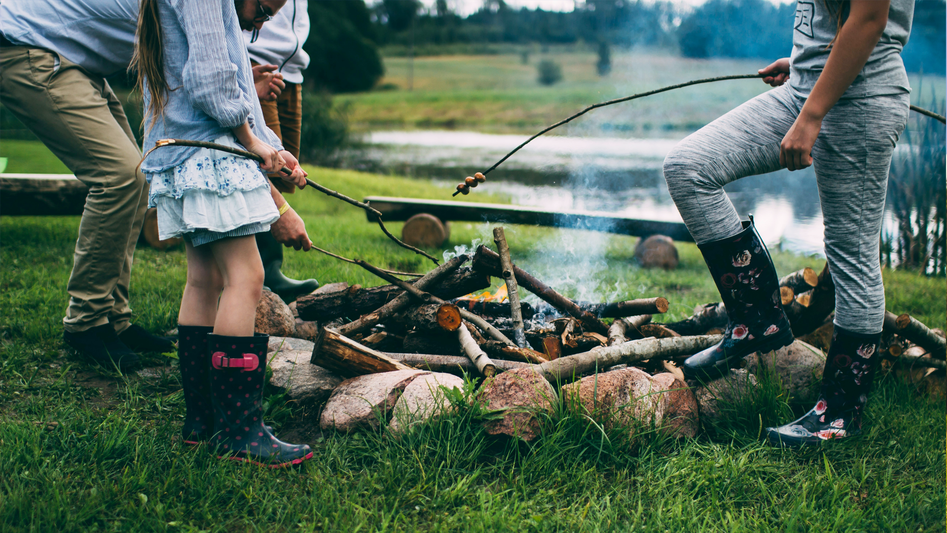 People gathered around a campfire by a lake, roasting marshmallows on sticks while wearing rain boots, with trees and water in the background.