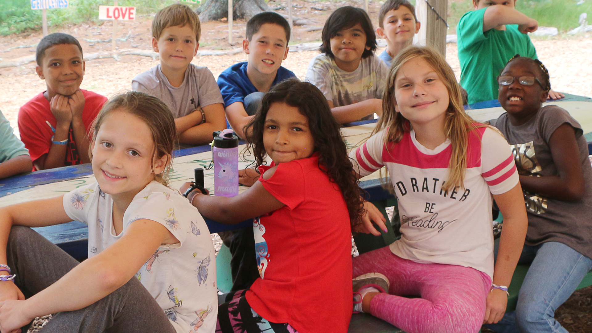 A group of children sitting at a colorful outdoor table, smiling at the camera, with a backdrop of trees and a sign that reads 'LOVE'.
