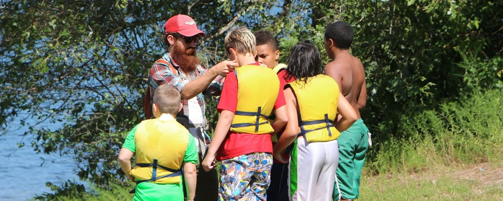 Group of children and an adult wearing life jackets beside a lake with trees in the background, possibly preparing for water activities.