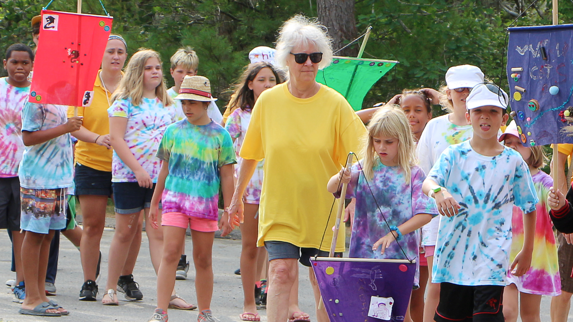 Group of children and a woman walking outdoors, some children wearing tie-dye shirts and hats, some holding colorful flags, in a wooded area.