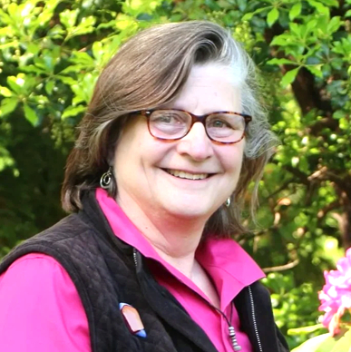 A smiling woman with short gray hair wearing glasses, a pink shirt, and a black vest standing outdoors among green trees and pink flowers.