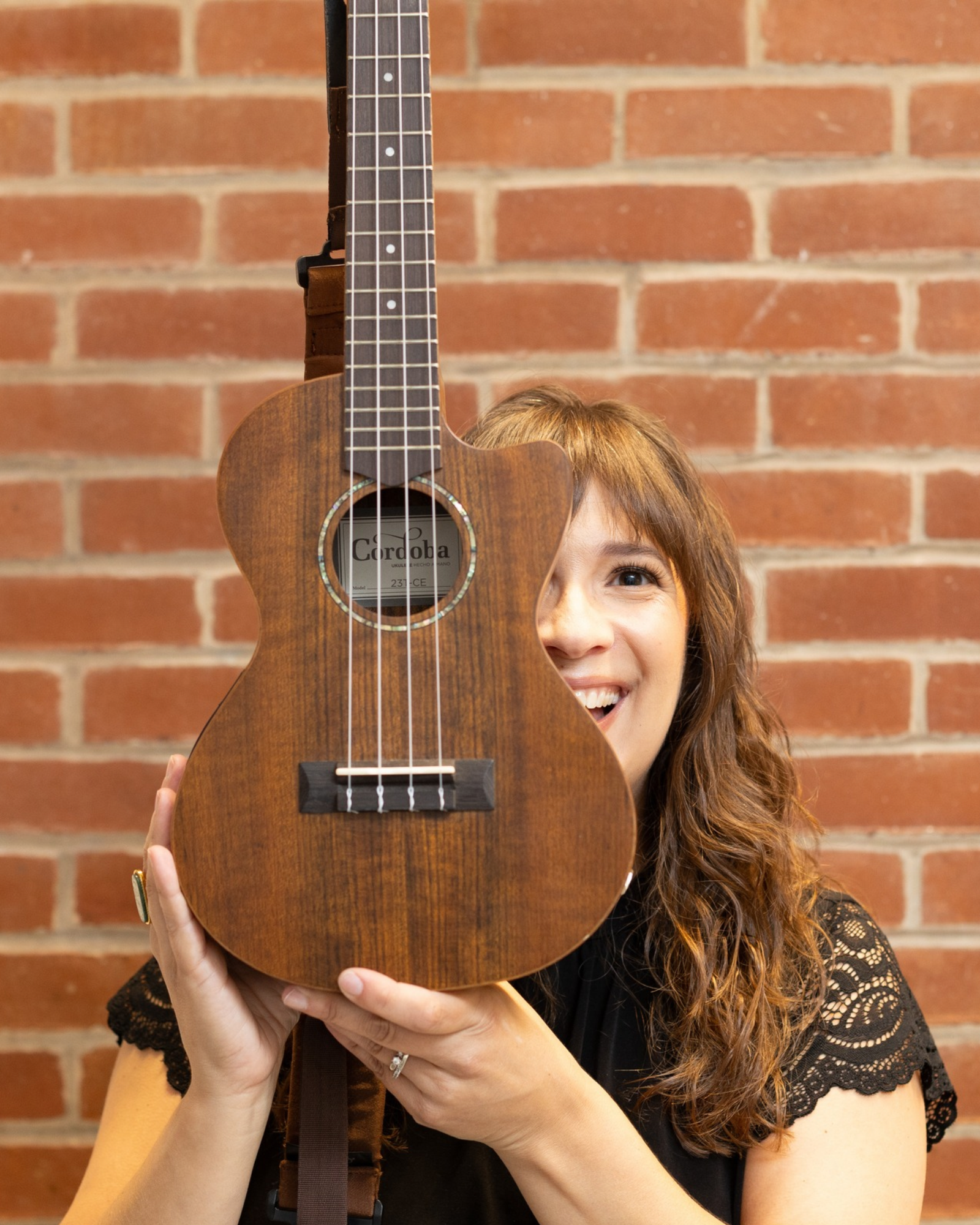 Sarah Keely Decker smiling from behind a wooden ukulele in front of a red brick wall.