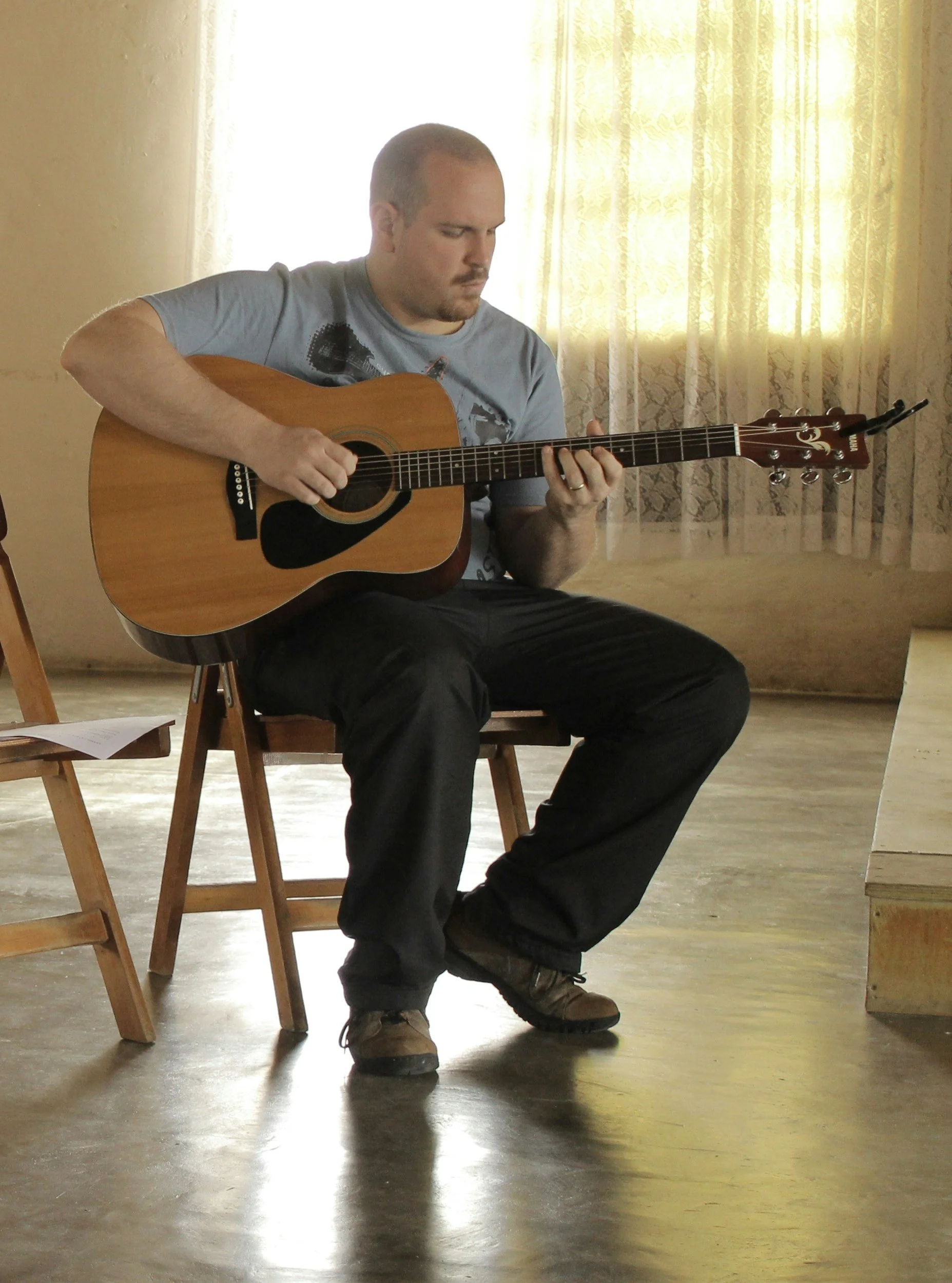 A man sitting on a wooden chair playing an acoustic guitar in a room with yellow curtains and wooden flooring.