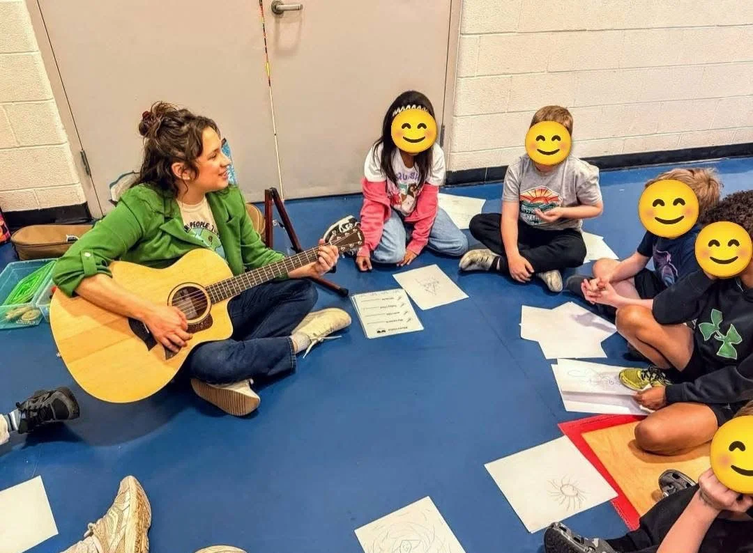 Sarah Keely Decker wearing a green jacket sits on the floor playing an acoustic guitar surrounded by five children with emoji faces drawn over their faces, sitting on a blue gym mat with drawings and papers in front of them.