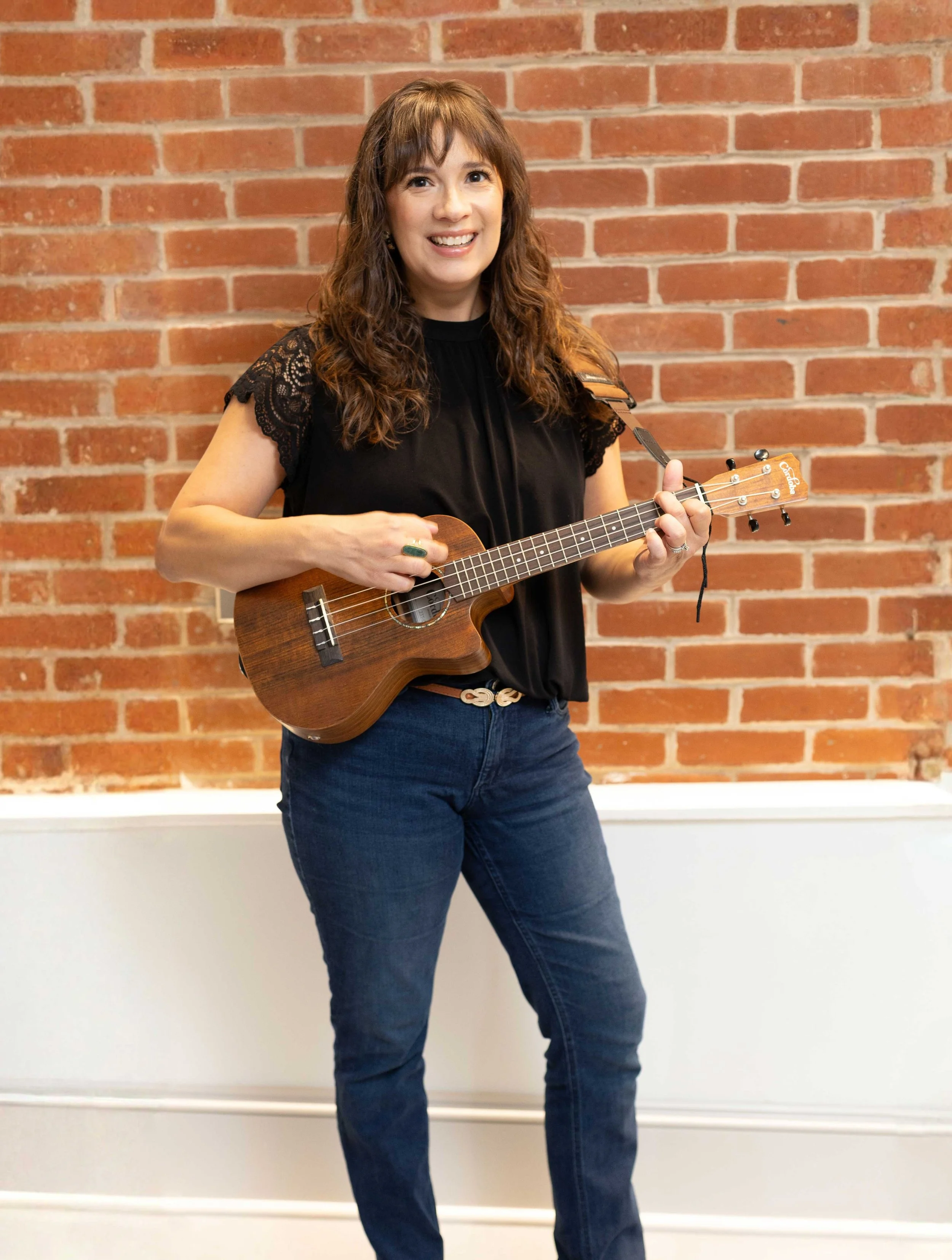 Sarah Keely Decker smiling while holding a ukulele in front of a red brick wall.