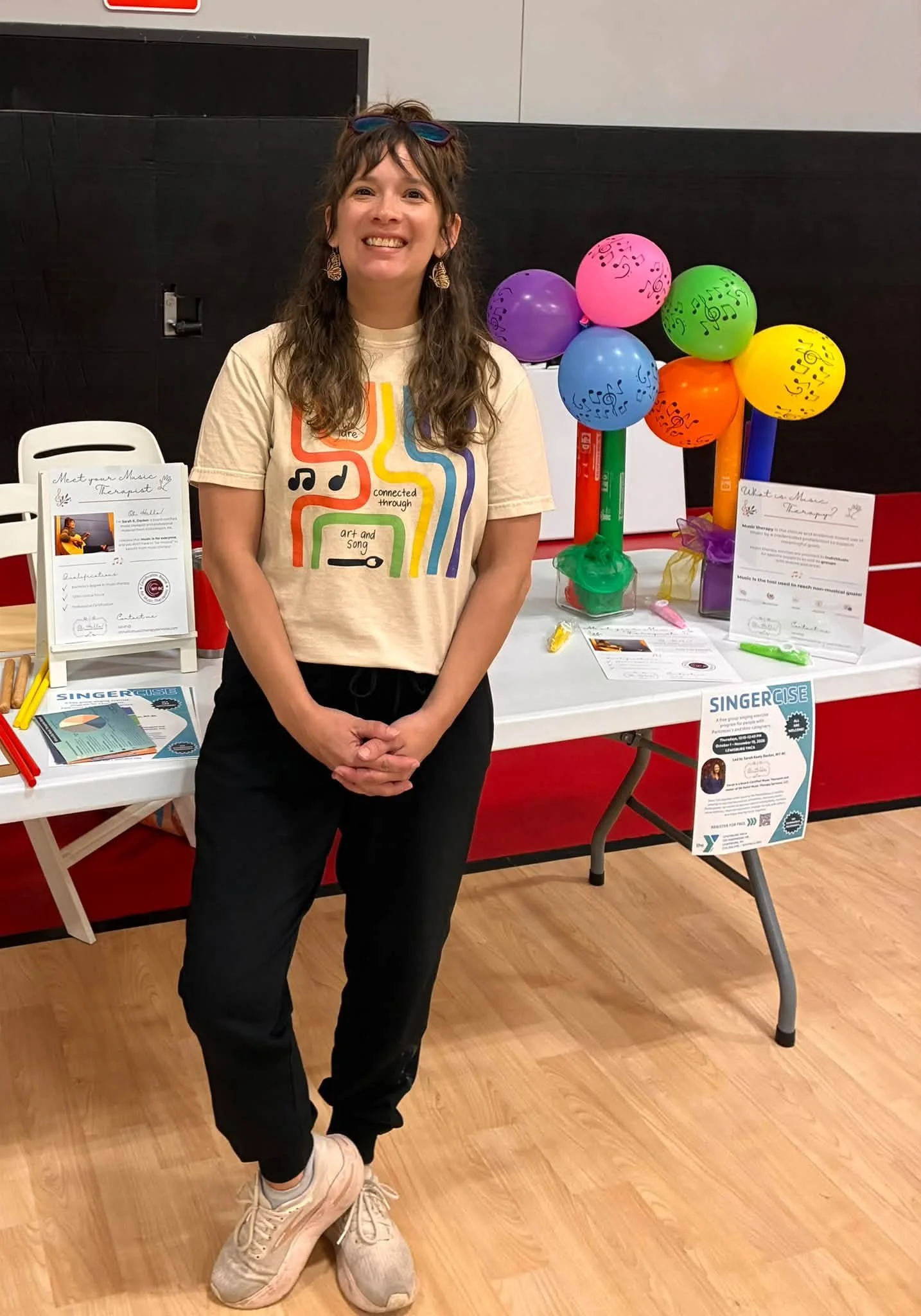 Sarah Keely Decker smiling stands in front of a table with colorful balloons and informational posters at an indoor event.