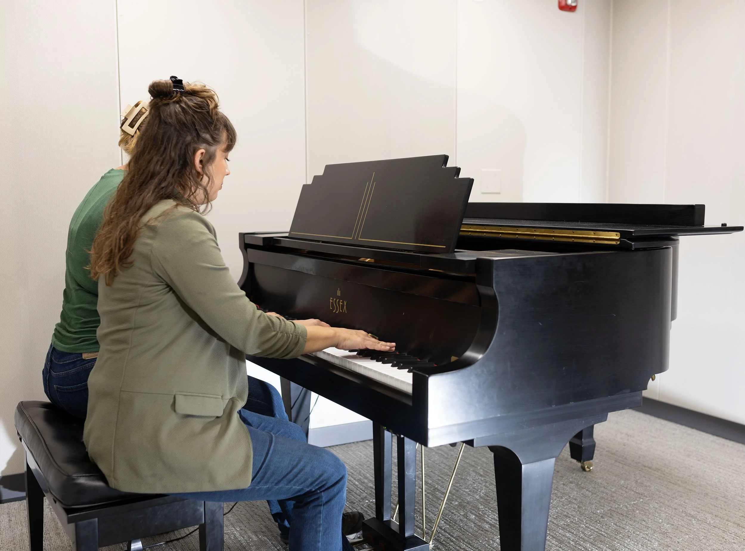 Sarah Keely Decker sitting on a piano bench, playing a black grand piano inside a room with white walls.