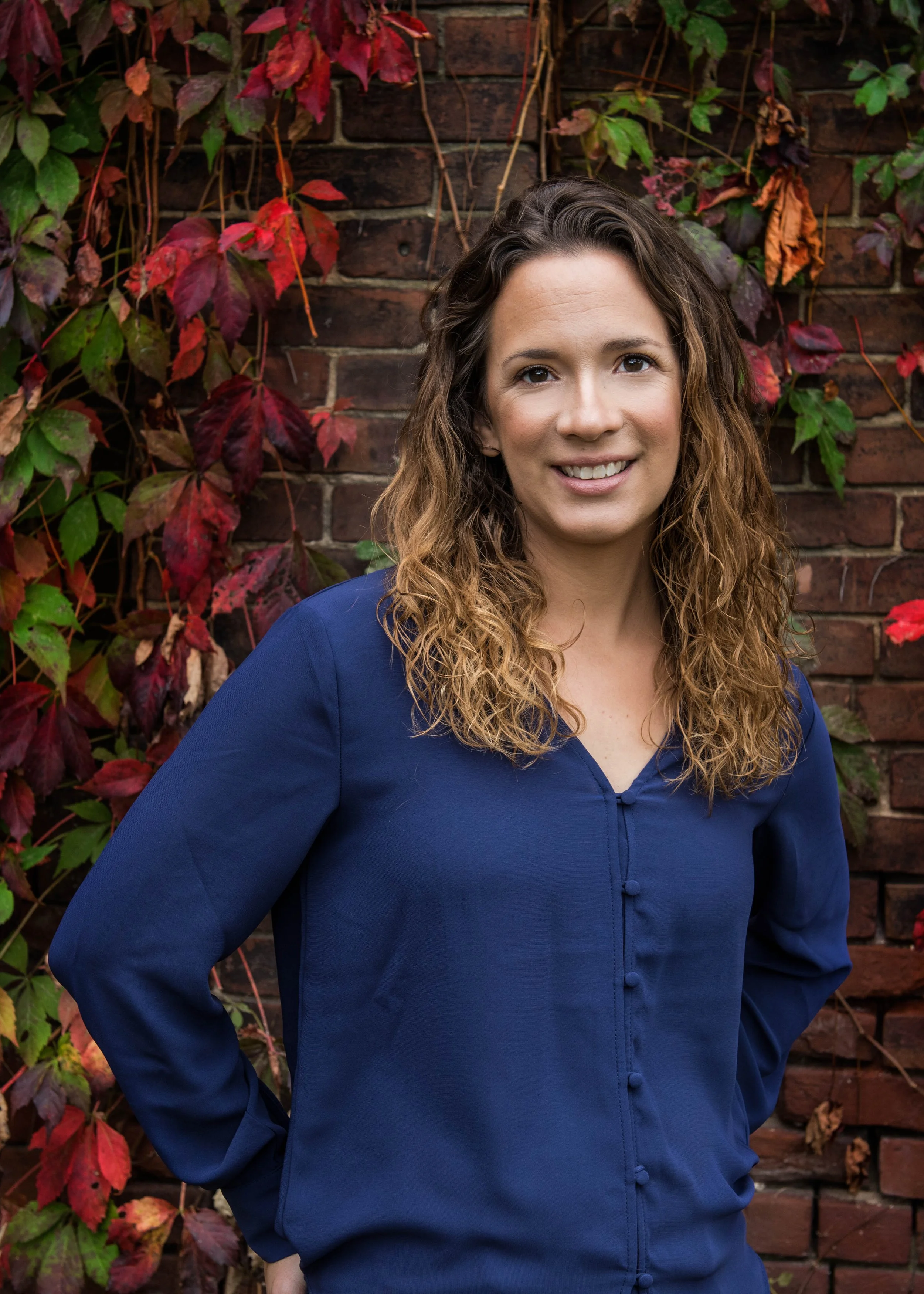 Sarah Keely Decker smiling in a navy blue blouse in front of a brick wall covered with red and green ivy.