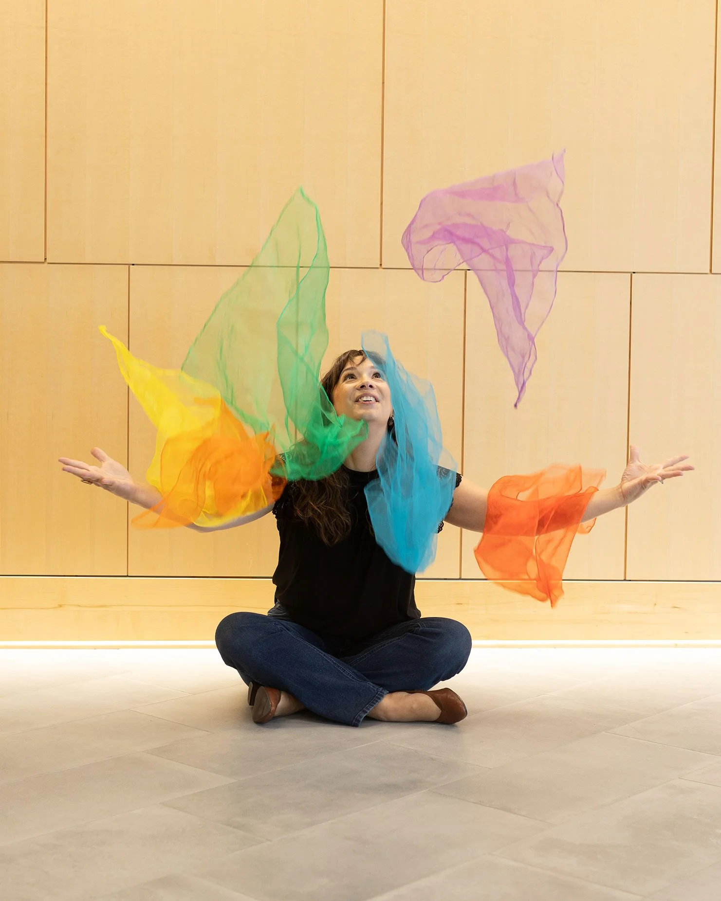 Sarah Keely Decker sitting cross-legged on the floor, surrounded by colorful sheer fabric, smiling and looking up, with a wooden panel wall in the background.