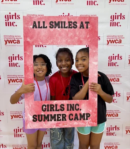 Three young girls smiling and holding a pink frame with text at a YWCA event promoting girls' summer camp in Nashville & Middle Tennessee.