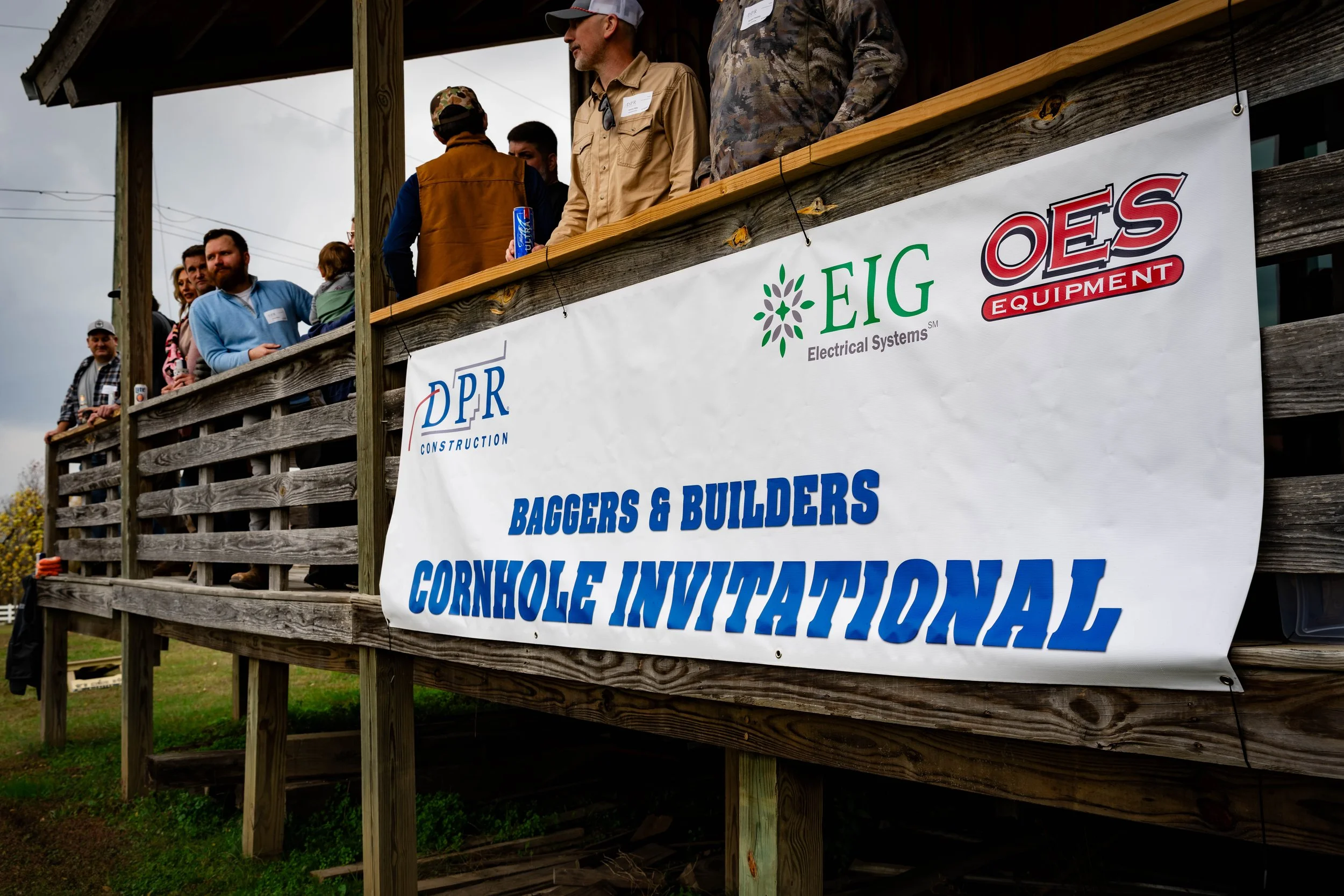 People standing on a wooden balcony at an outdoor event, with banners displaying logos for electrical systems, OES Equipment, and a cornhole tournament.