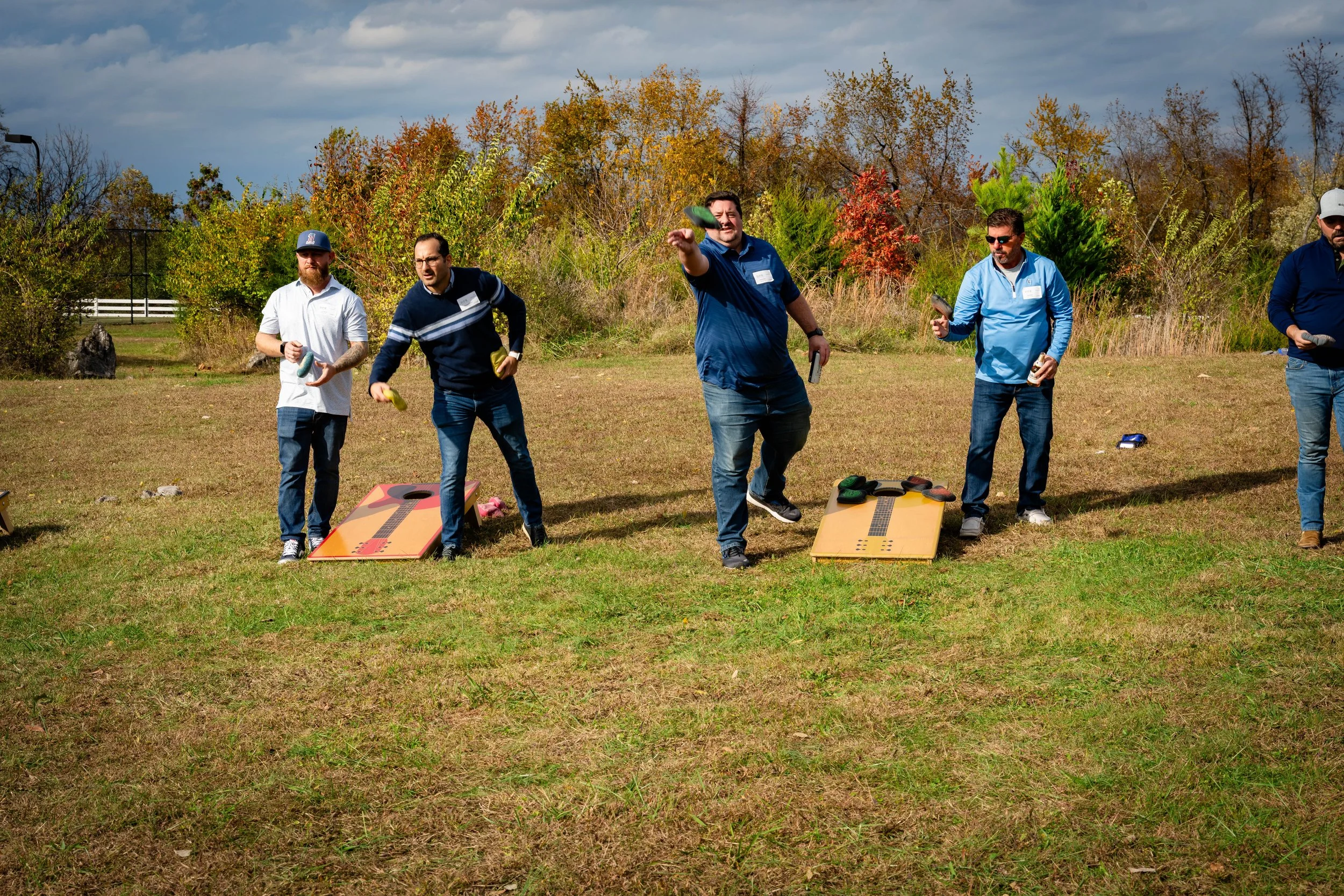 A group of five men playing cornhole outdoors on a grassy field with trees and a cloudy sky in the background.