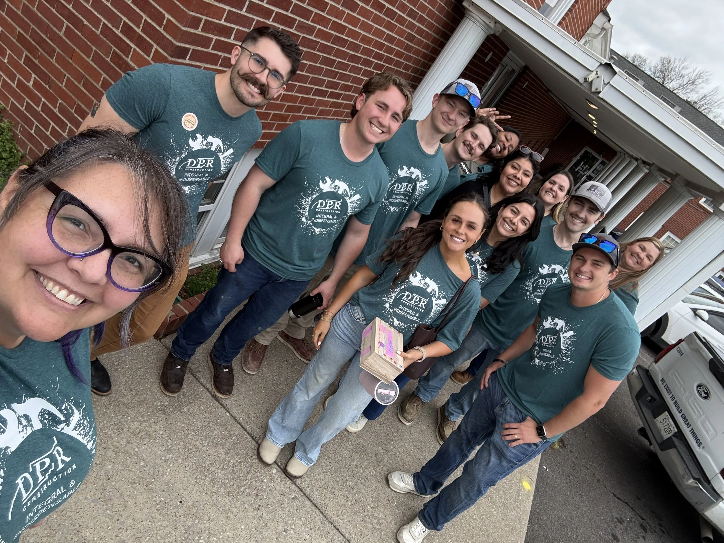 A group of smiling people standing in front of a brick building, wearing matching teal T-shirts with a circular logo that says 'DPR Construction' and 'Integral & Indispensable.' They are gathered outdoors, with some holding items and one person wearing sunglasses on her head. The group appears to be participating in a community or team event.