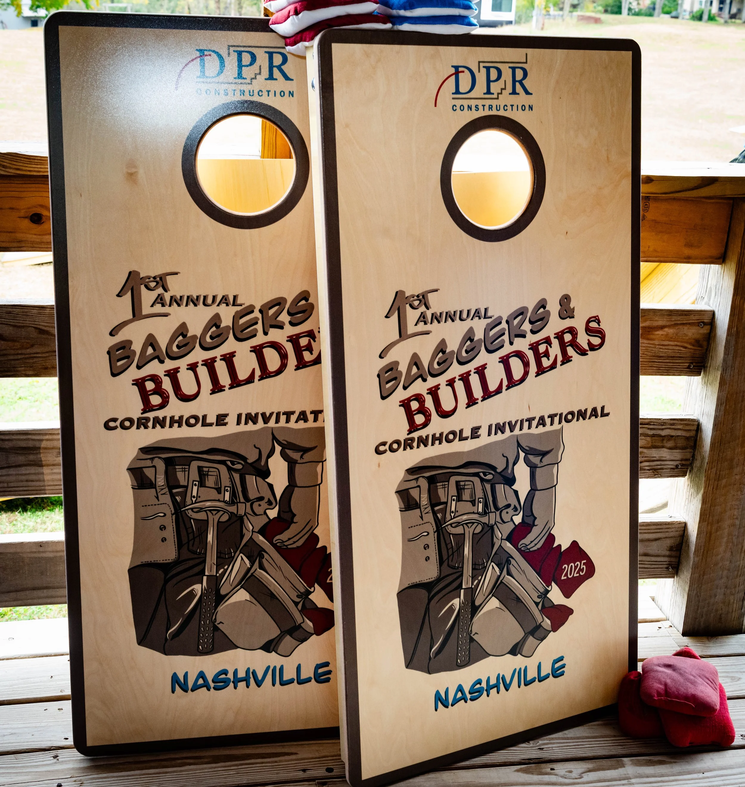 Two cornhole boards with a Nashville theme lean against a wooden railing, with a pink bag nearby. The boards feature a graphic of a person throwing a beanbag and text advertising an annual event, with the year 2025 visible.