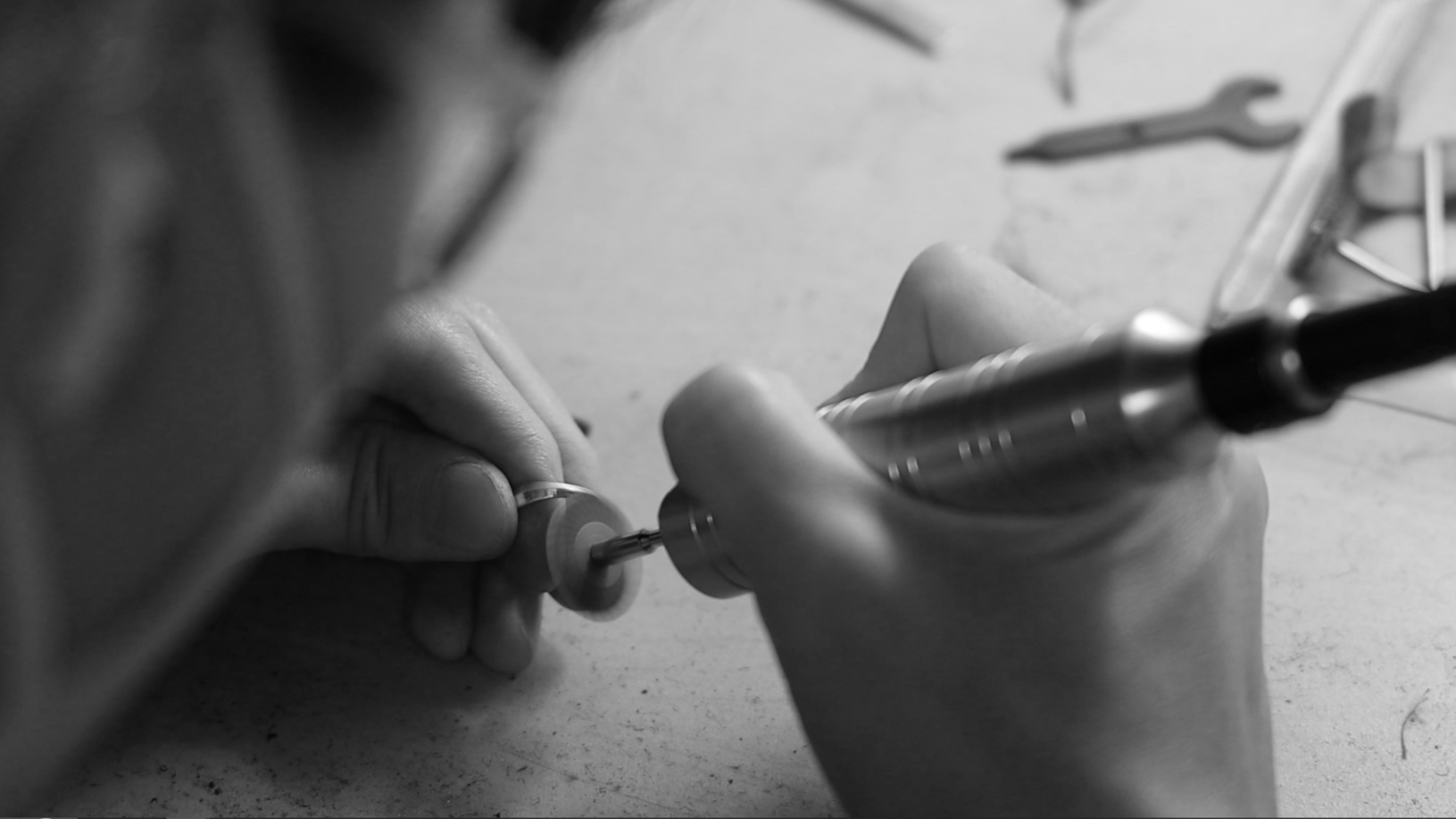 Close-up of a person working with a small drum on a workbench, using a rotary tool to shape or engrave it, with various tools and equipment in the background.