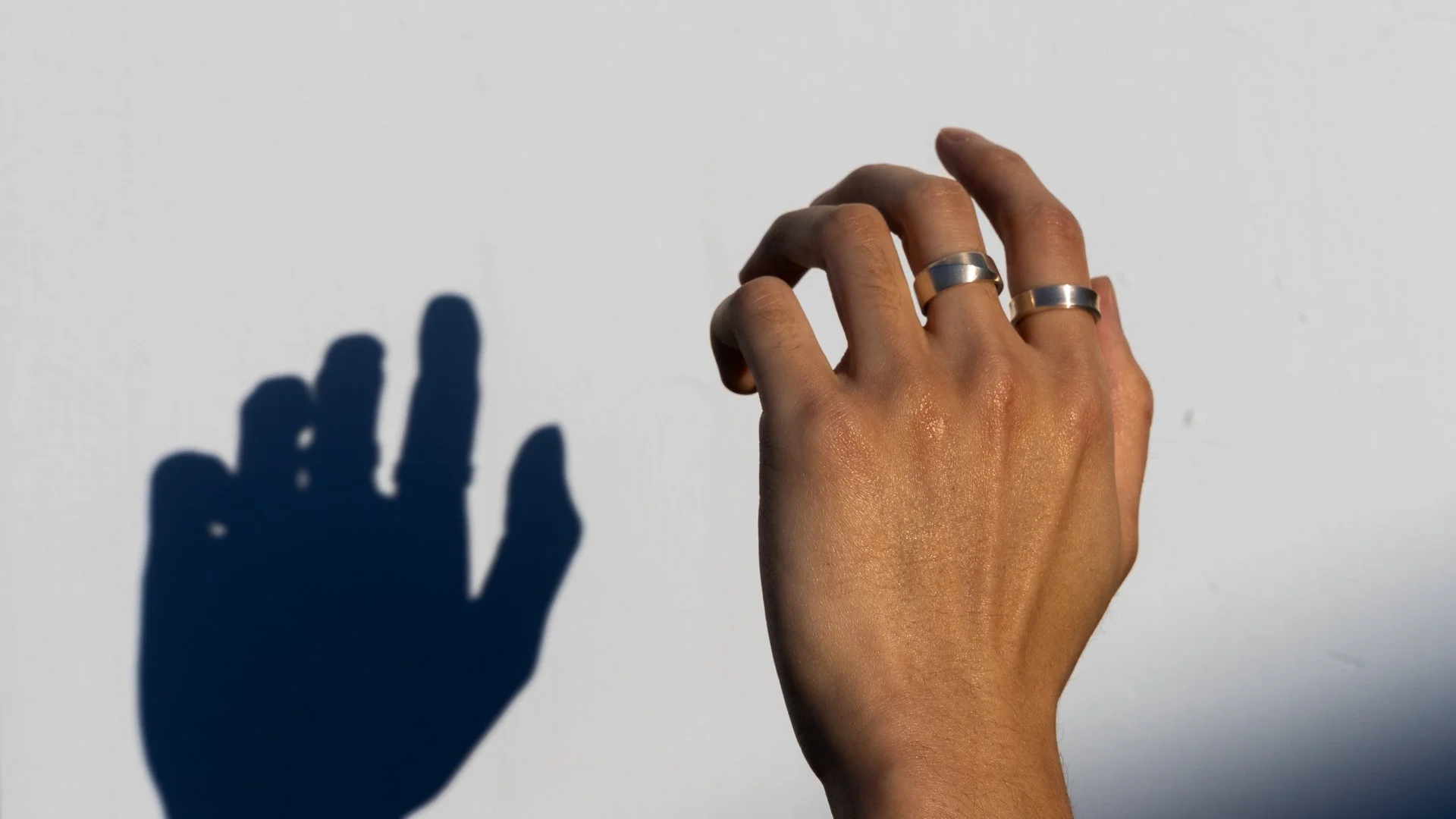 Close-up of a person’s hand with two rings on fingers, reaching toward a white wall to cast a shadow resembling a hand with spread fingers.
