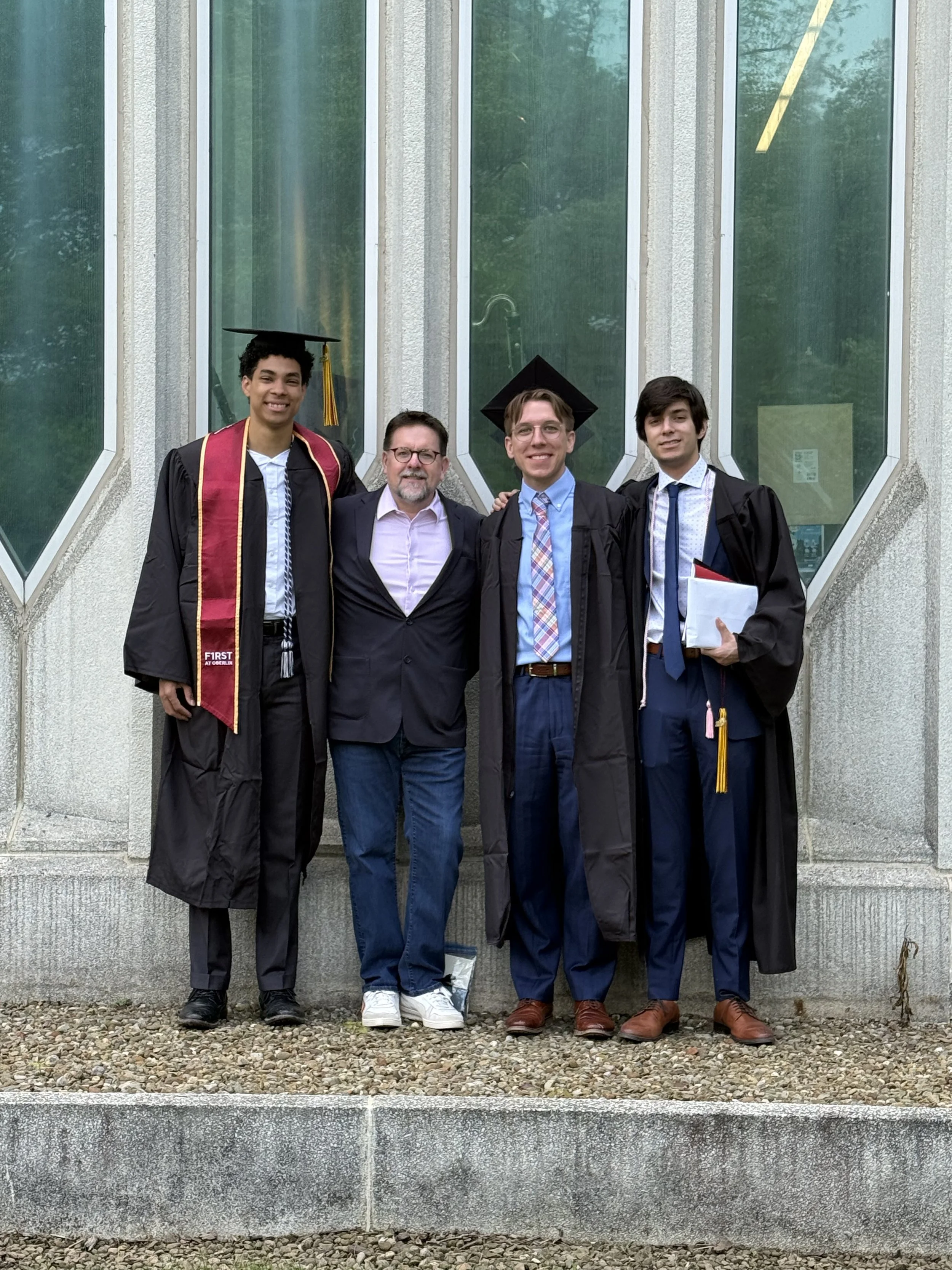 Four people wearing graduation gowns and caps, standing outdoors in front of a building with large green-tinted windows. They are smiling, and some are holding folders or documents.