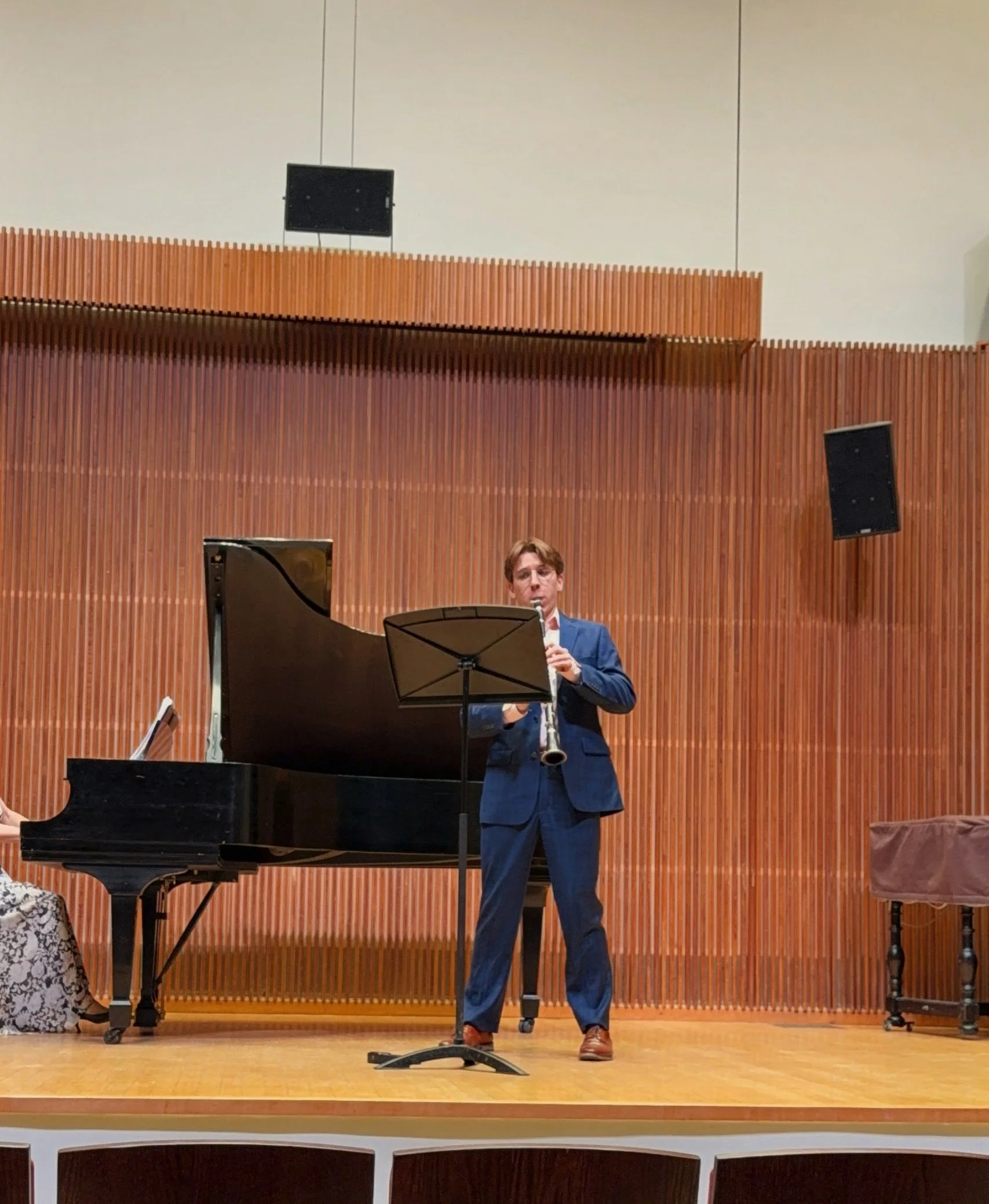A man in a blue suit playing a clarinet on a stage with wooden paneling, a grand piano, and a black music stand, with black speakers mounted on either side.
