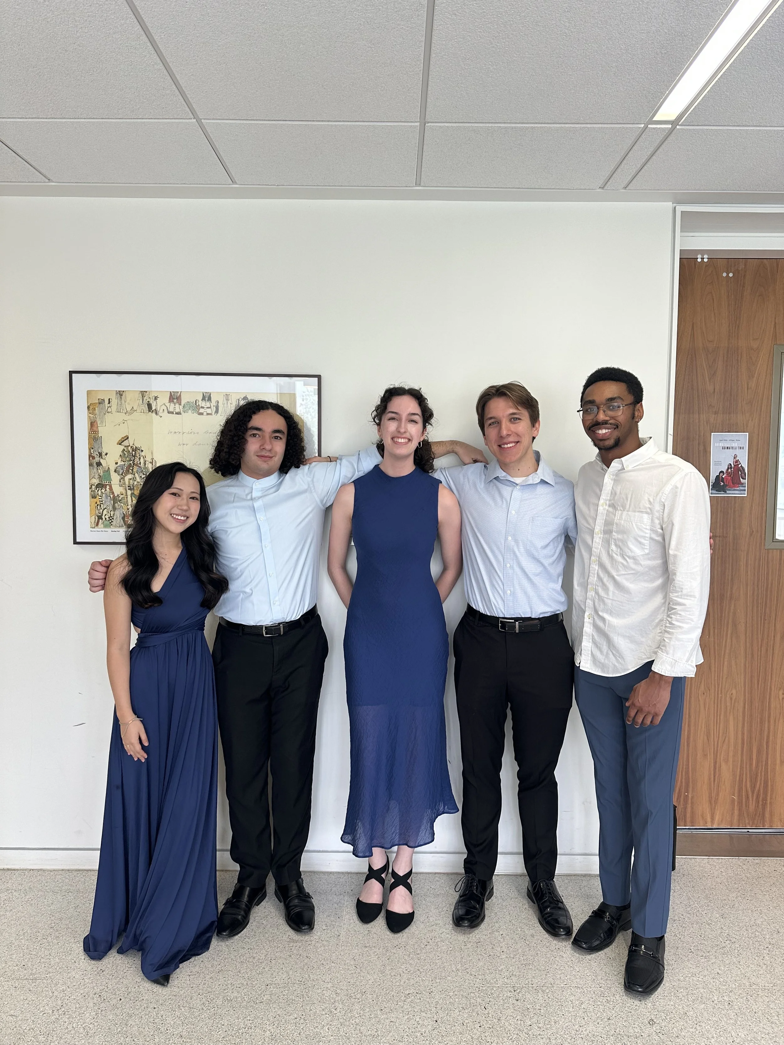 Group of five young adults standing together indoors, dressed in formal attire, smiling at the camera.