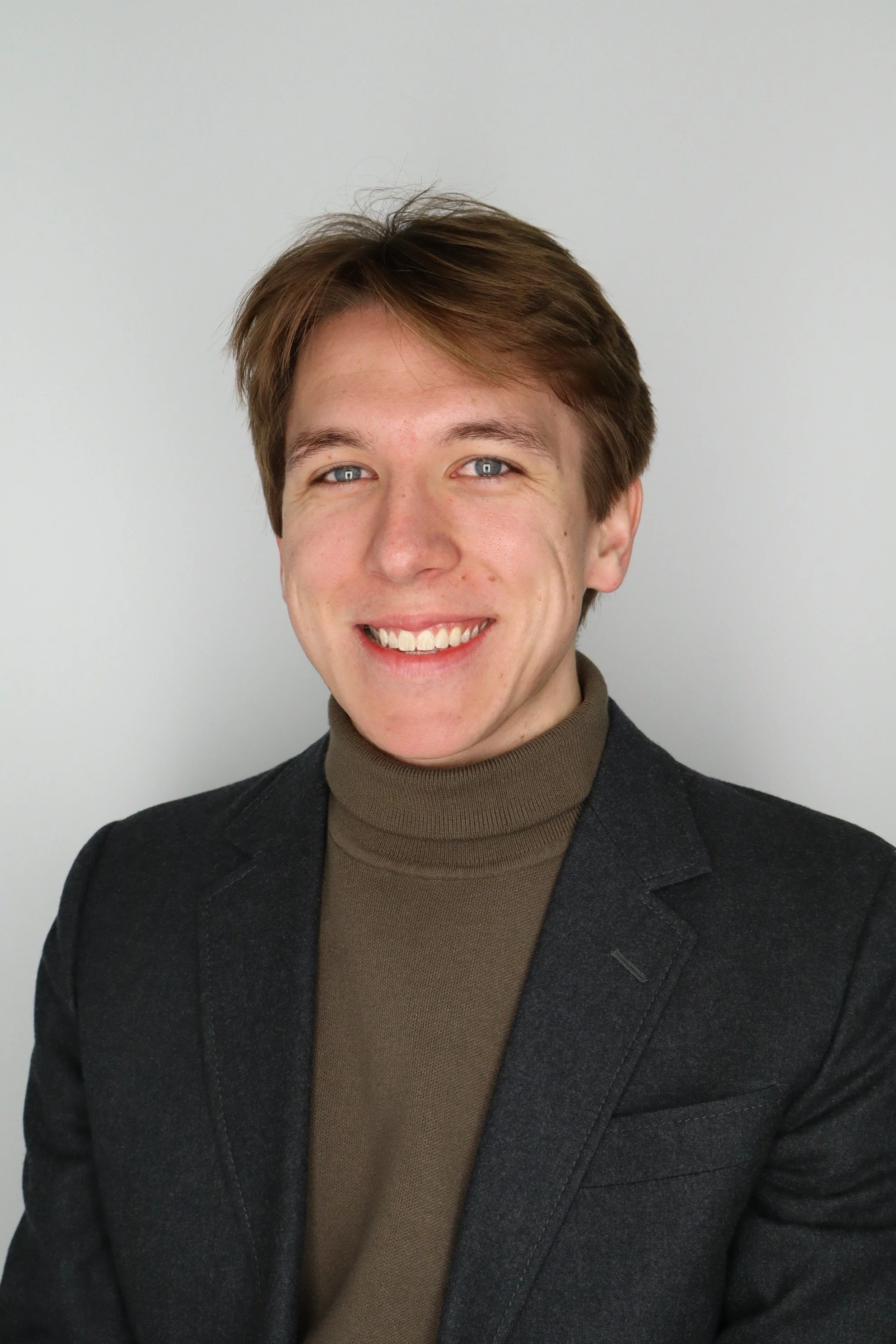 A young man with light brown hair styled to the side, blue eyes, and a bright smile, wearing a dark blazer over a brown turtleneck sweater, standing against a plain light gray background.