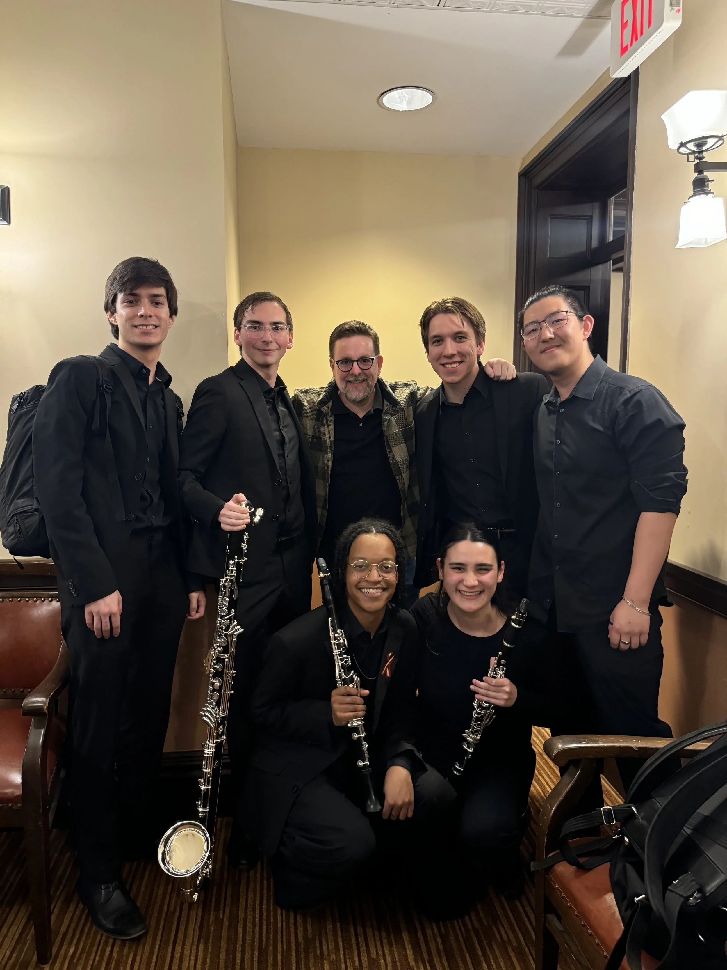 Group photo of six people, five men and one woman, posing with musical instruments including clarinets and a flute in a room with yellow walls, wooden furniture, and an emergency exit sign on the ceiling.