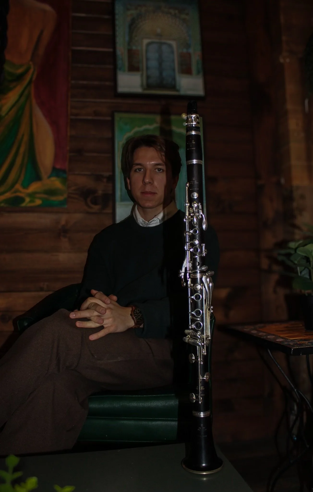 A man sitting with folded hands, wearing a dark sweater and a watch, holding a black clarinet with silver keys, in a wooden room with paintings on the wall.